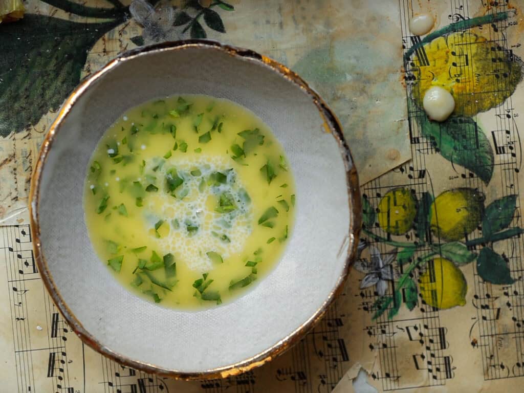 A bowl of vegan egg wash mixture with chopped herbs on a vintage sheet music page featuring lemon illustrations. The edges of the bowl are gold-rimmed, and the surface below includes splashes of the mixture.